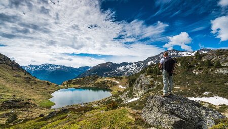 A hiker stands with their back to the camera on a rock while gazing over a small lake, with mountains in the background