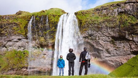 Family of travellers look at Seljalandsfoss waterfall as a rainbow forms in the mist in the Golden Circle of Iceland