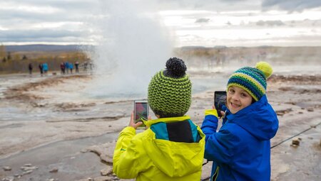 Young travellers smile and watch as Strokkur geyser erupts in front of them as they film on their phones