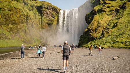 Intrepid traveller walks up to Skogafoss waterfall at the heigh of Iceland summer with numerous other travellers beyond