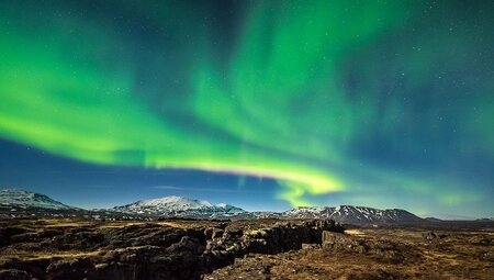 Green waves of the Northern Lights undulate over the rocky mountainous landscape of Thingvellir National Park, Iceland