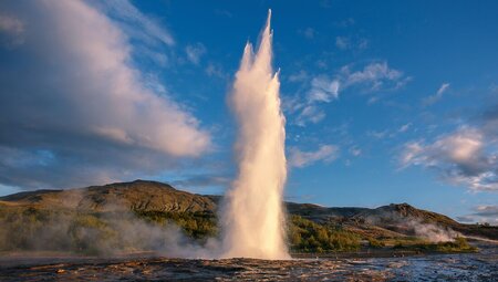 Strokkur Geyser erupting during sunset with blue sky and white fluffy clouds