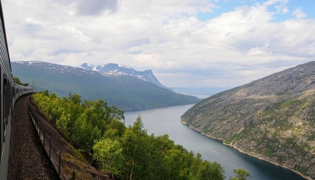 A train rides a high ridge along the Otenfjord with a forest below and mountains looming beyond