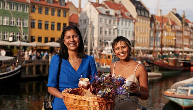 Two 18 to 35 travellers smile at the camera holding ice cream and a bike on the canal in Copenhagen, Denmark