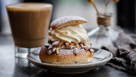 An appetizing Semla bun with almonds and heavy cream, flour and sugar and a hot latte behind