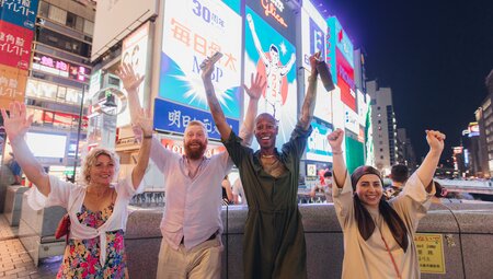 Intrepid travellers throw their hands up surrounded by screens and lights in Dotombori