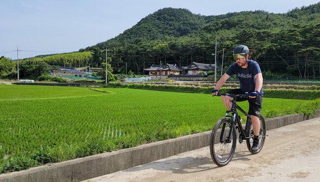 Cyclist in helmet and gear cycling in rural South Korea past traditional houses and fields