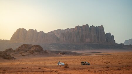 Spire mountain range in distance with two trucks driving past in the distance in an orange sand desert