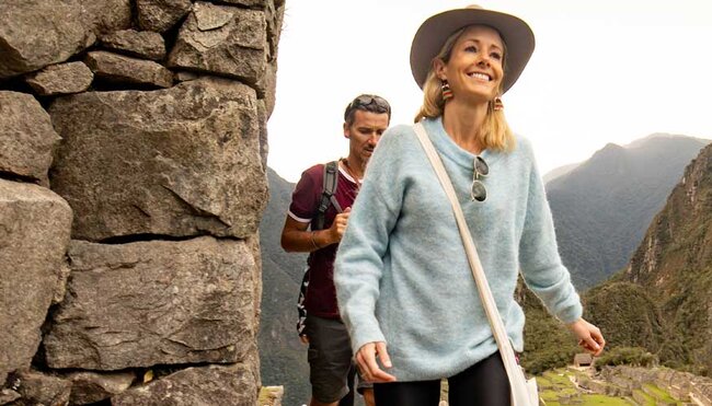 A blonde woman with a blue sweater walk in front of a man in a red t-shirt, with Machu Picchu in the background