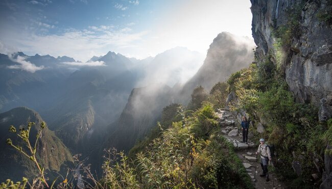 Travellers look back at the photographer as sunlight hits a gentle mist around the bend of a mountain path on the Inca Trail