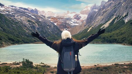 Traveller stands with outstretched hands overlooking green Laguuna Esmerelda with mountains in the background