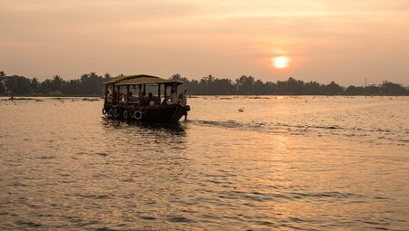 Small cruise ship sails across a lake in the Kerala Backwaters with a red orange sunset in the background