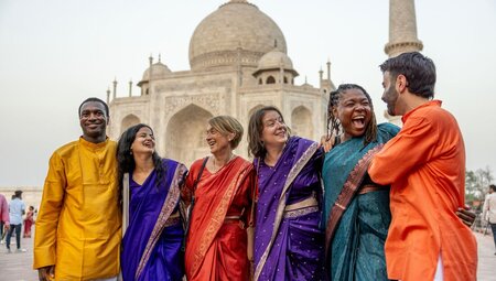 Travellers in opulent colourful garments smile and laugh happily together outside the Taj Mahal at sunset
