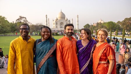 Group of varied ages pose with smiles in colourful elegant clothing standing at a distance in front of the Taj Mahal