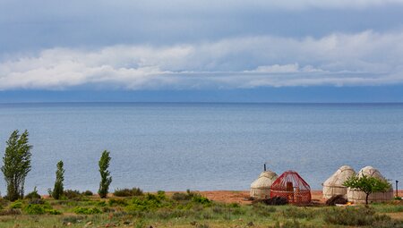 A set of yurts with one partially assembled sit on above the shore of Issy-Kul Lake with mountains in the distance