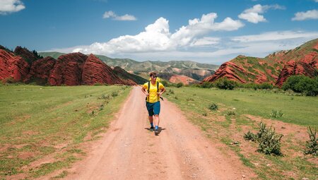 Traevller in yellow hikes in the red and green landscape of Jeti Oguz with clear sunny clouds in background