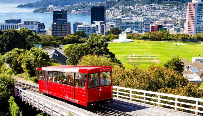 Red tram on tracks in Wellington, with cityscape and a cricket field in the background, New Zealand