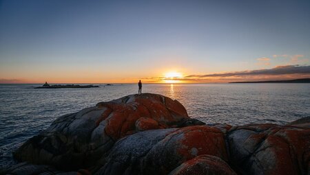 Intrepid traveller stands on Bay of Fires boulder shore looking out at the sunset over the Tasman Sea