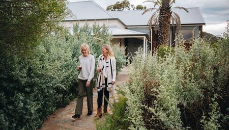 Travellers smiling and enjoying a walk with drinks on the accomodation grounds with green plants around them, Arkaba, South Australia