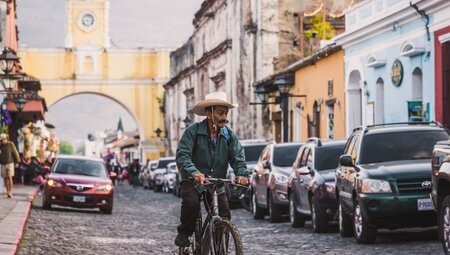 Local man in wide brimmed hat riding a mouuntain bike down the streets of Antigua with old buildings and new cars around