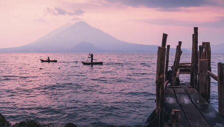 Locals on boats paddle across the reflective surface of Lake Atitlan at sunrise with volcano beyo9nd