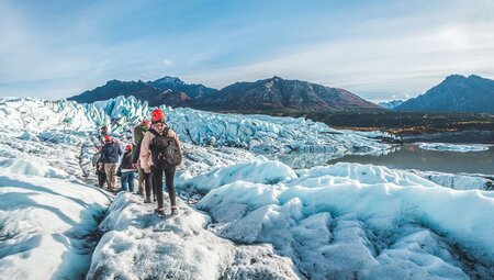 Column of travellers in safety gear hike on the icey Matanuska Glacier with Alaskan landscape in the distance