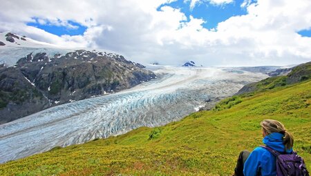 Blonde female traveller sits on a hill overlooking Exit Glacier in Harding Ice Field