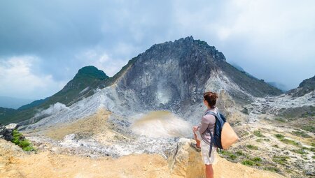 Traveller looks out at caldera emitting heat and steam at top of Mt Sibayak in Sumatra from end of a hike