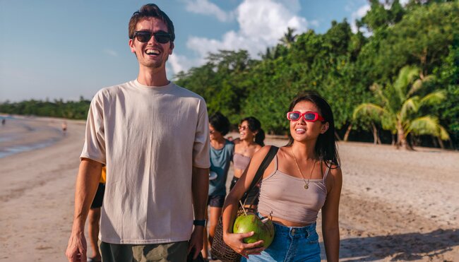 Young Intrepid travellers stroll along a beach in the Philippines with drinking coconuts laughing and enjoyng the sun