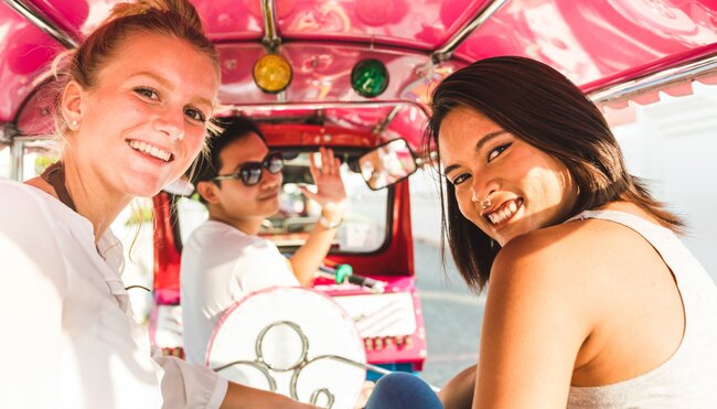 Friends smile at camera enjoying a tuk tuk ride in downtown Bangkok in Thailand