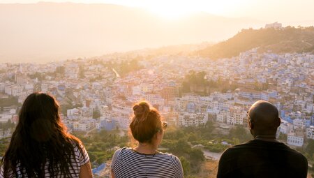 Three travellers sit on a high overlook looking out over blue painted buildings of Chefchaouen