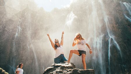 Travellers stand on rock under the spray of waterfall