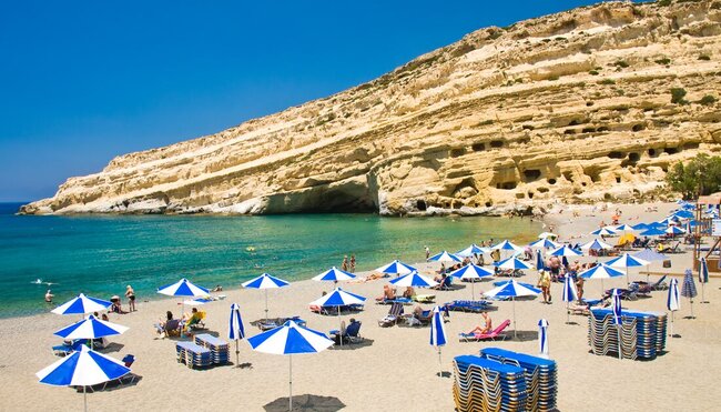 Travellers on the beach with blue and white parasols Matala beach with cliff in background, Crete, Greece