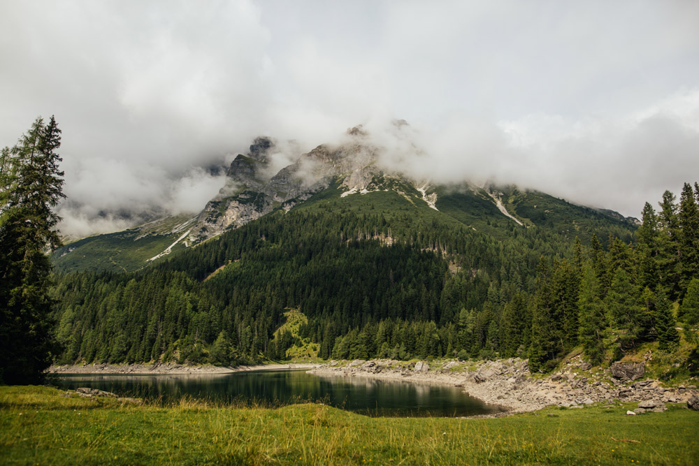 kleiner see, hinter dem sich mit wald bewachsene berghänge befinden, wolkenverhangener himmel