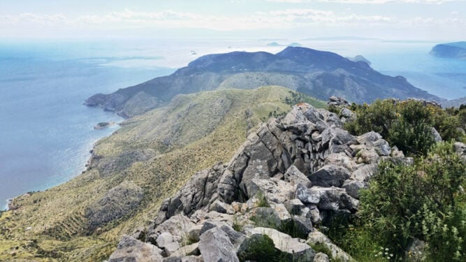 Blick von felsigem Gipfel hinunter ans Meer, grüne Flächen und wenige Wolken am Horizont