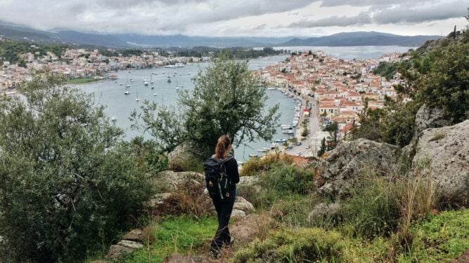 Wanderin steht auf einer Anhöhe mit Aussicht auf eine Stadt an einer Küste, bewölktes Wetter