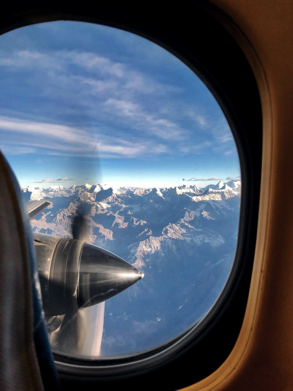 Blick aus Bullauge von Flugzeug auf teilweise schneebedeckte Berge. Blauer Himmel, wenige Wolken.