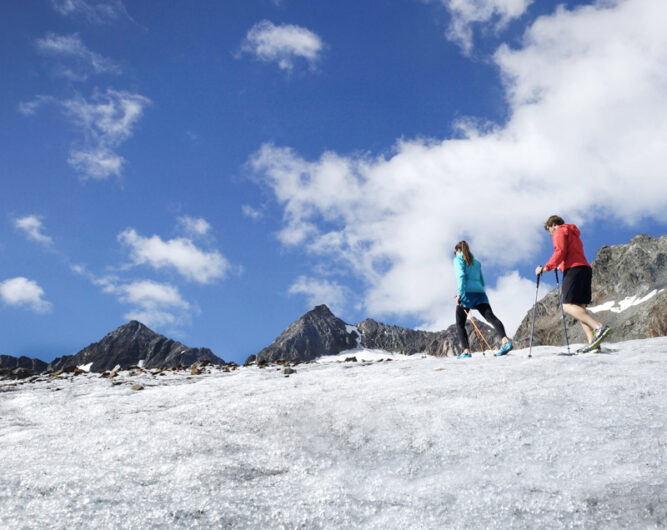 Zwei Bergsteiger auf einem Schneefeld. Die vordere Person in blauer, die hintere in roter Jacke. Blauer Himmel