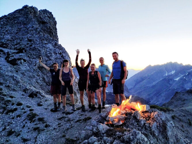 Sechs Personen auf einem Berg, vor ihnen ein Feuer. Abend