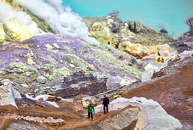 Zwei Männer tragen Körbe voller Schwefe von einer Mine aus auf einen Berg.