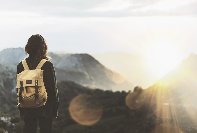 Frau mit Rucksack von hinten vor Bergkulisse im Sonnenuntergang