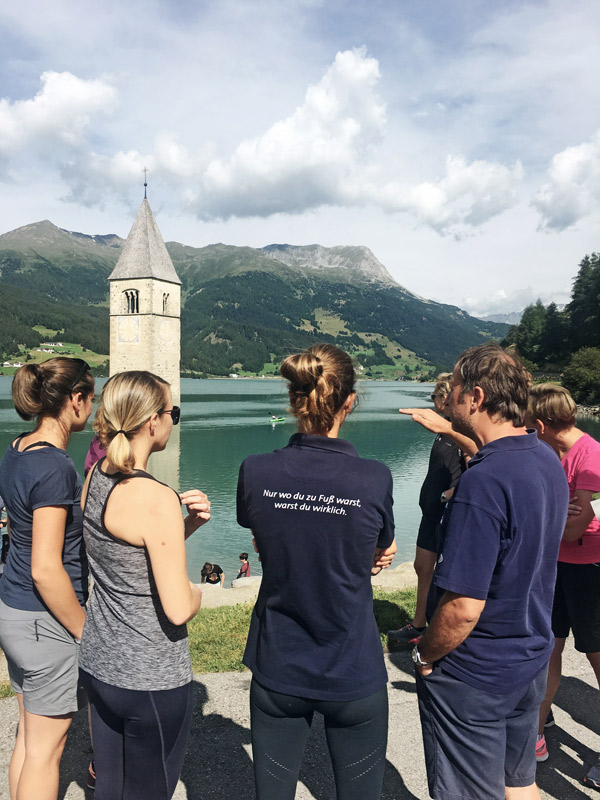 Gruppe von Menschen vor einem See mit einem Kirchturm im Wasser.