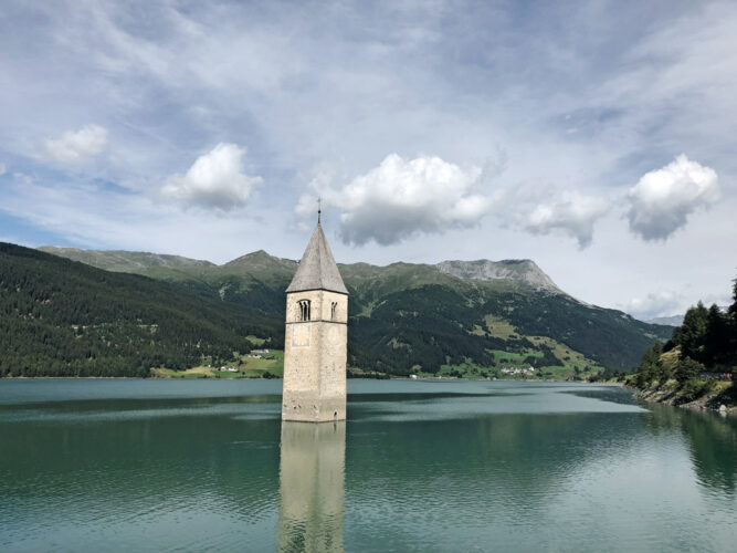 Kirchturm in einem See, dunkelgrünes Wasser, Wolken am Himmel