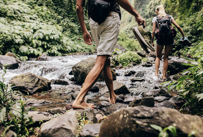 Mann und Frau, die im Wald mit den Schuhen in der Hand den Bach barfuß überqueren.