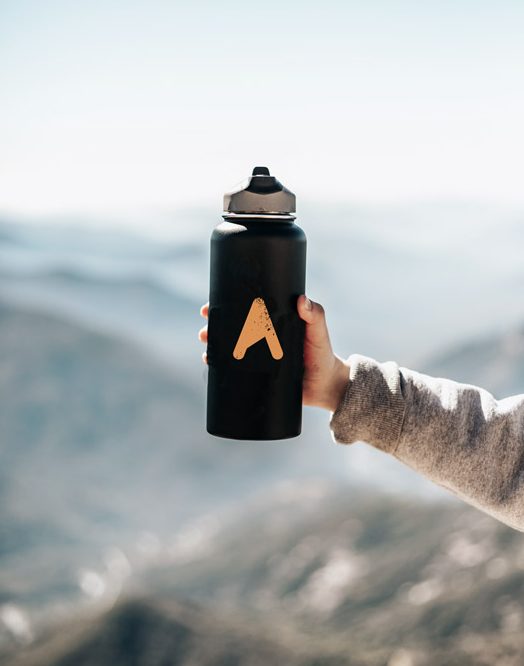 Eine Wasserflasche mti dem ASI Logo in einer Hand. Im Hintergrund sind Gipfel zu sehen.