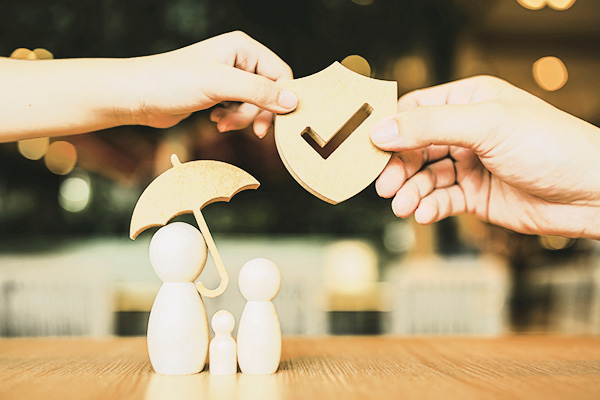drei Holzfiguren mit einem Schirm und zwei Händen darüber, die ein Schutzschild aus Holz halten