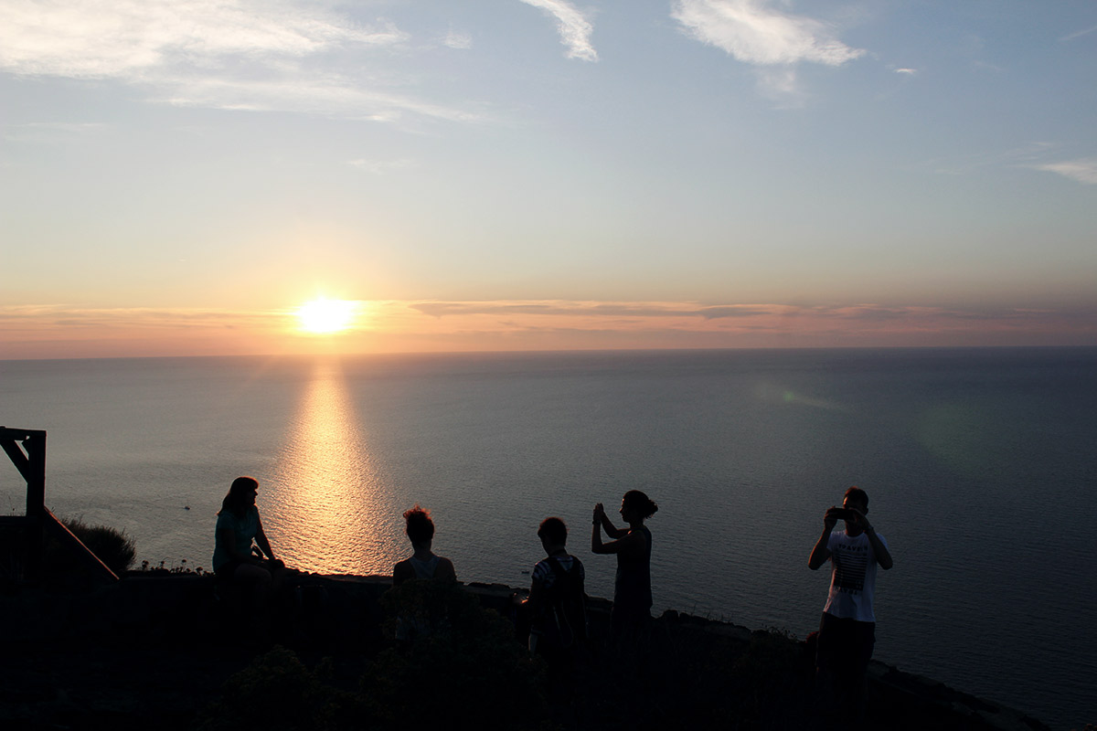 untergehende Sonne am Horizont, davor meer und silhouetten von menschen 