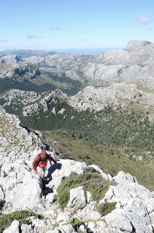 Wanderer in roter Jacke auf felsigem Bergsattel. Ausblick in die Ferne zu einem See und zur Küste. Blauer Himmel