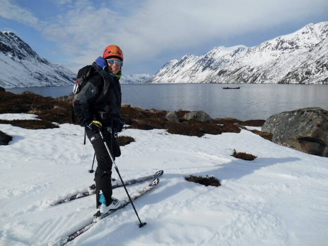 Skitourengeher lächelt in die Kamera, steht an einer verschneiten Bucht vor dunkelblauem Meer