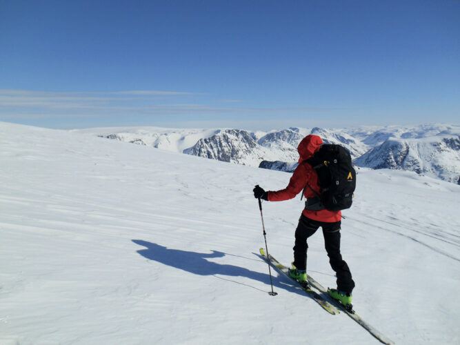 Ein Skitourengeher mit roter Jacke von hinten, weiße Landschaft und verschneite Berge im Hintergrund, klarer Himmel